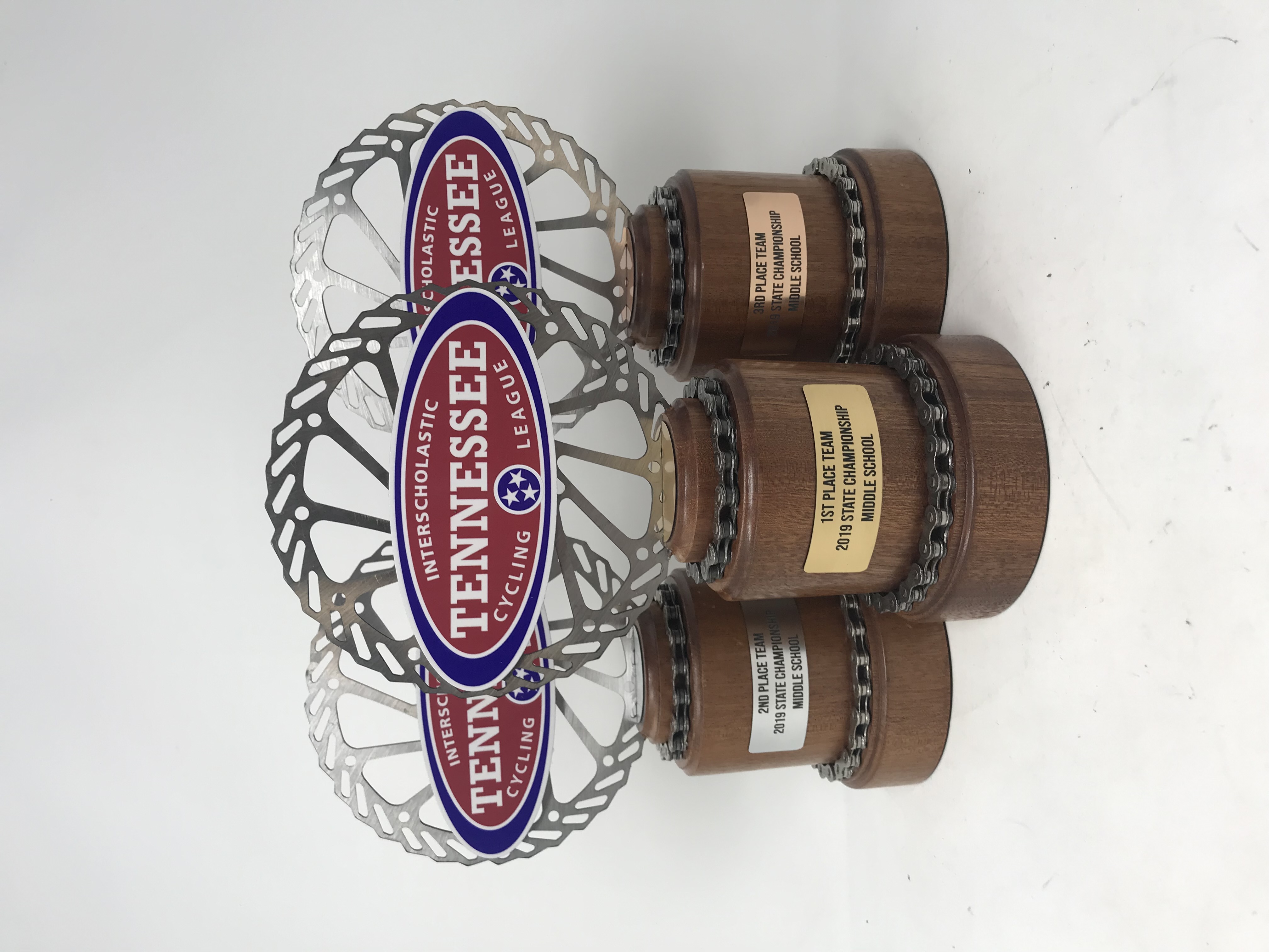 Three Noble Series trophies, hand-spun dark mahogany bases banded with reclaimed bike chain, each topped with a steel disc rotor carrying the Tennessee Interscholastic Cycling League crest and a brass team plate