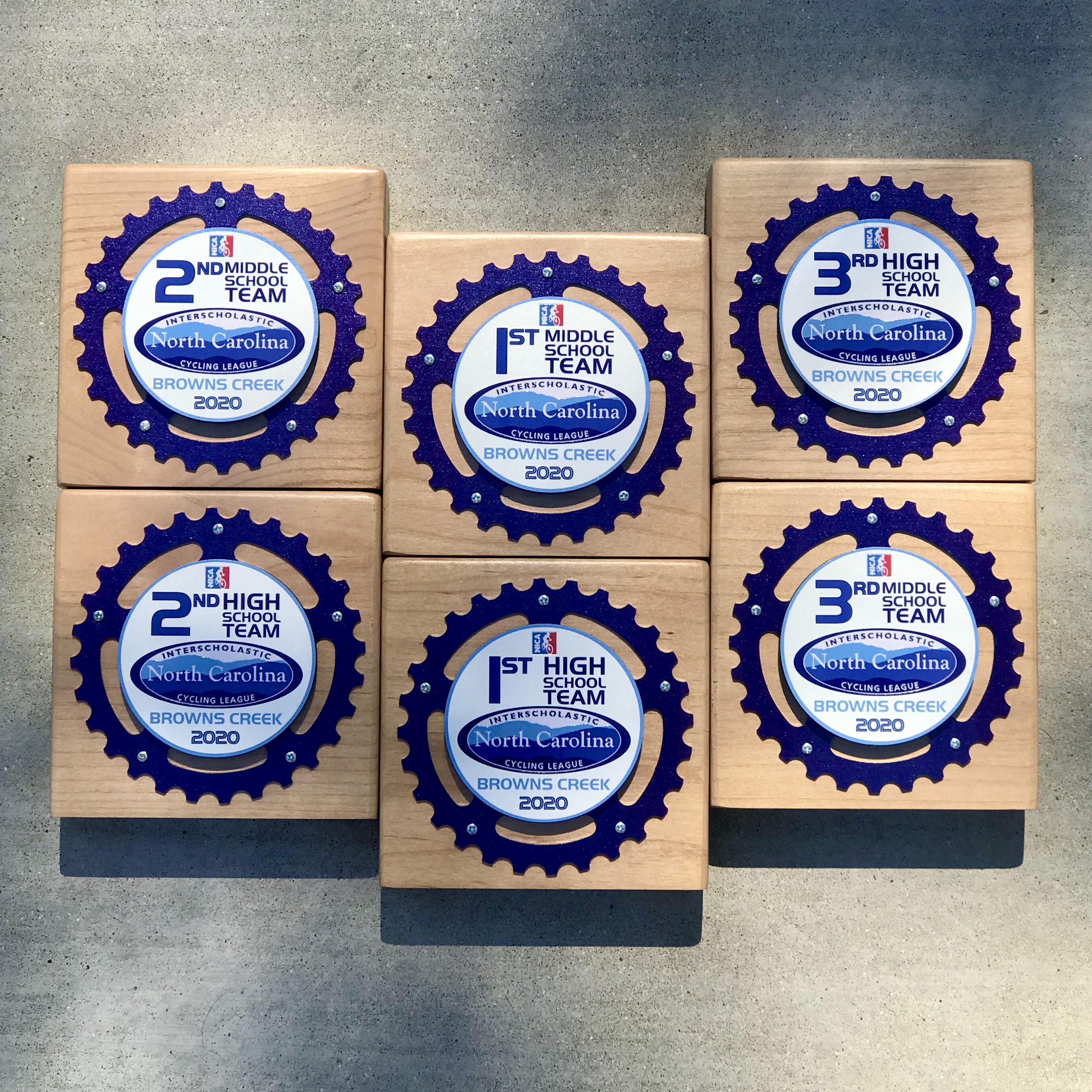 Six Triumph Series maple block awards for the North Carolina Interscholastic Cycling League at Browns Creek, each fronted with a purple gear ring around a printed 1st, 2nd or 3rd place middle school and high school team medallion