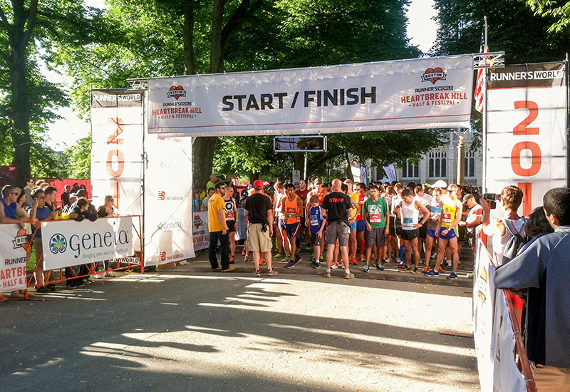 Runners massed under a START / FINISH vinyl banner on a scaffold gantry at the Heartbreak Hill Half and Festival, with sponsor banners along the barriers on both sides