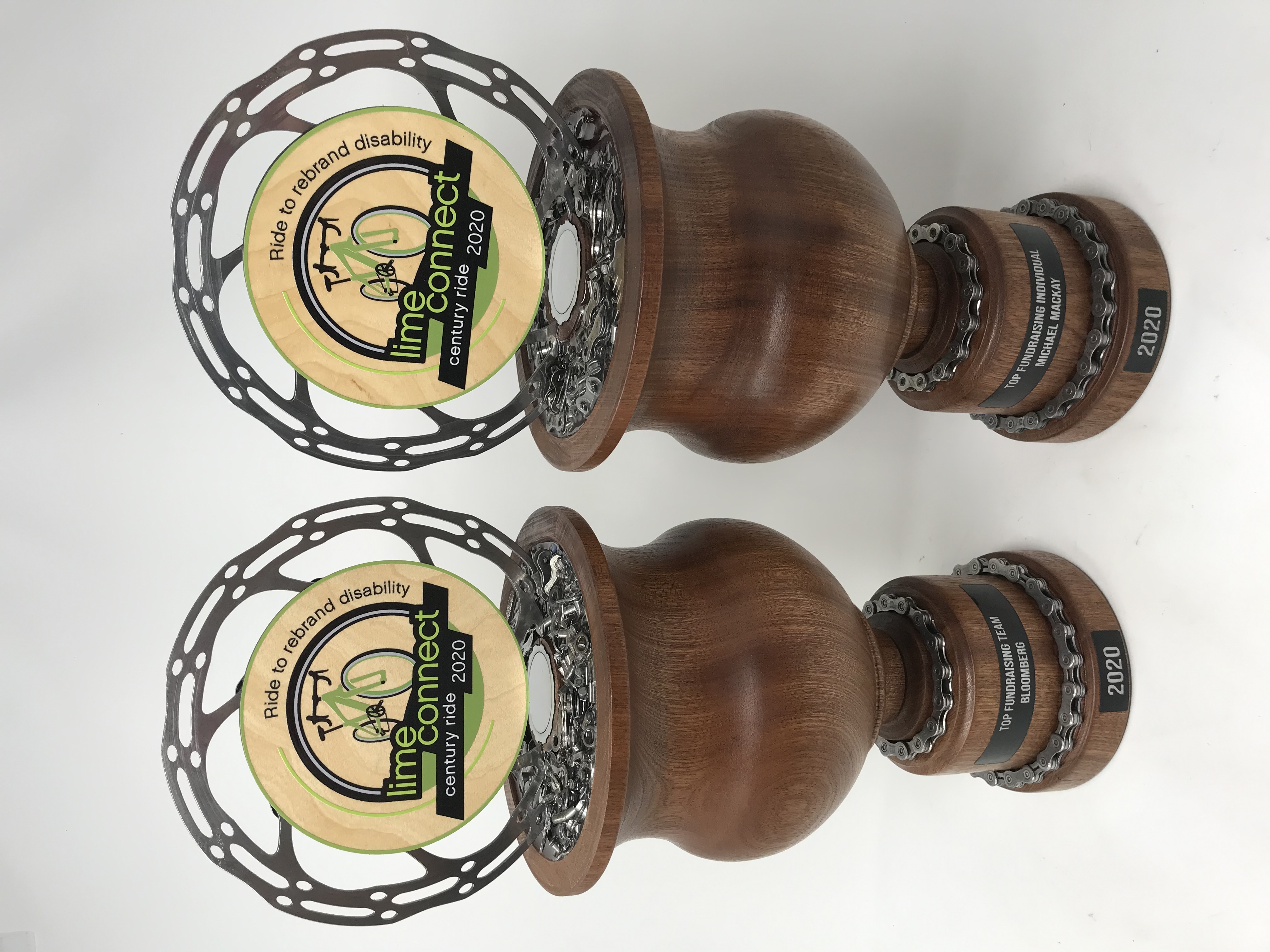 Two World Series trophies, turned walnut goblets banded with reclaimed chain, each mounted with a steel disc rotor carrying the Lime Connect century ride artwork and a chain-wrapped base plate