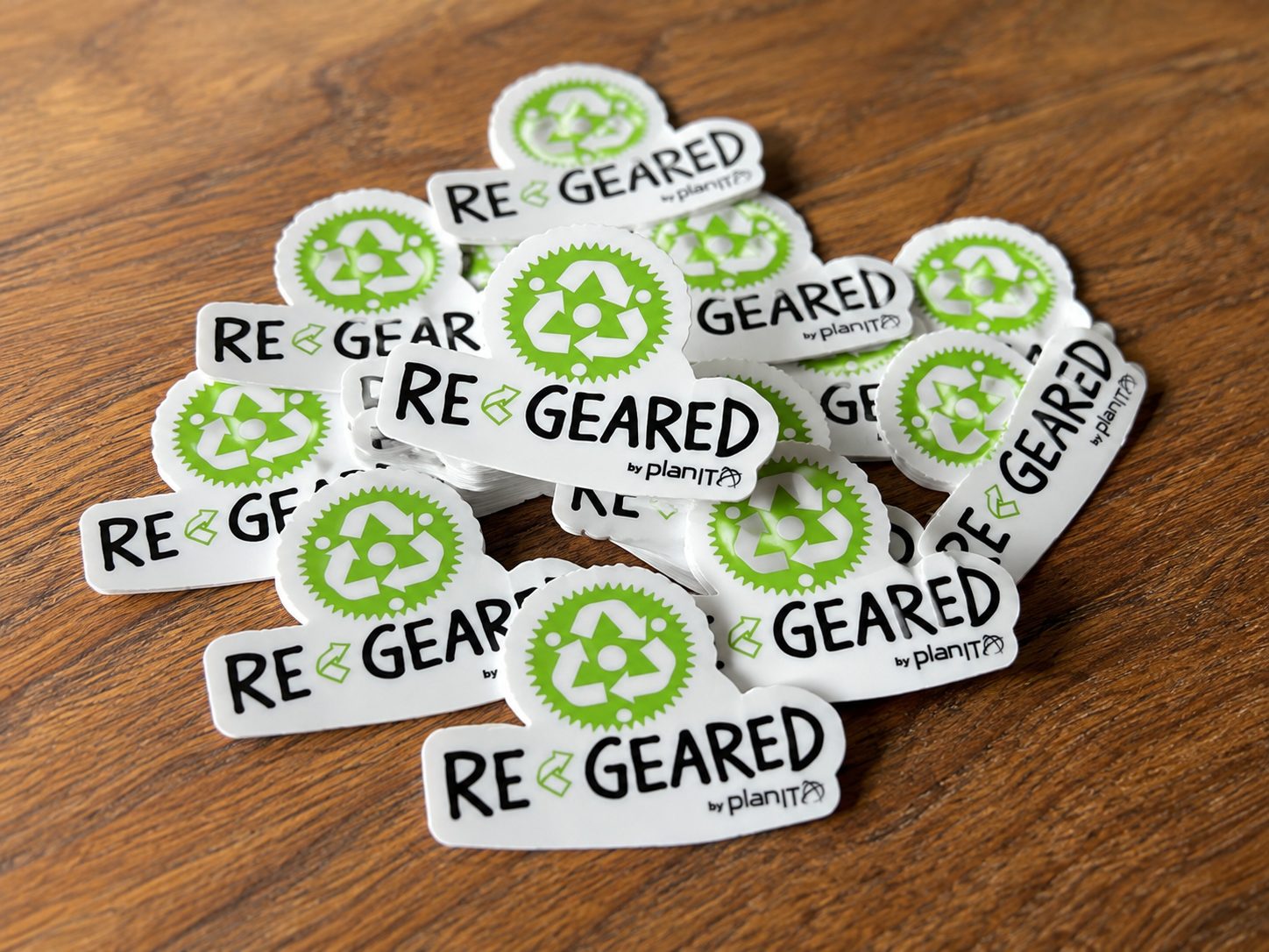 Pile of die-cut ReGeared stickers on a wooden table, each cut to the outline of the green recycled-chainring logo and black wordmark