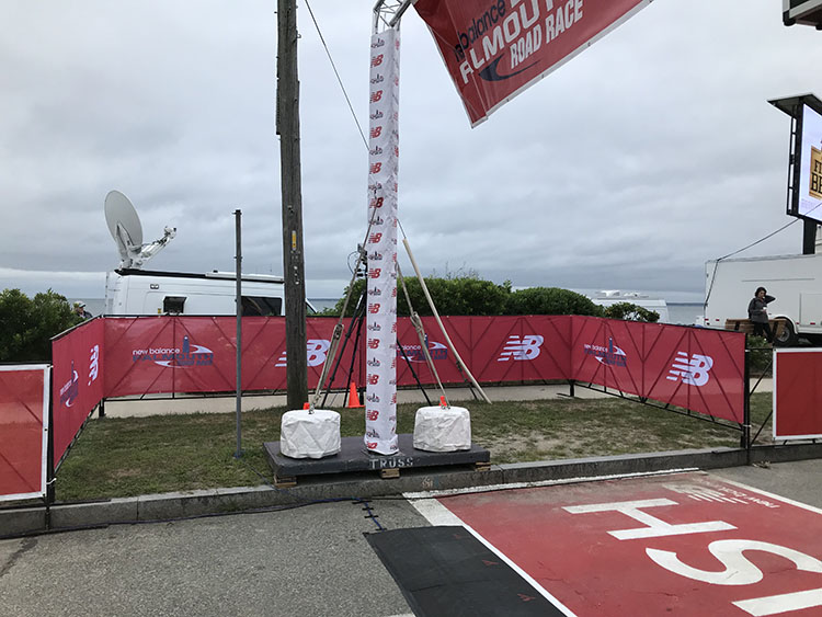 Red mesh lane control banners printed with New Balance branding, fenced along the roadside at the Falmouth Road Race beside a broadcast truck