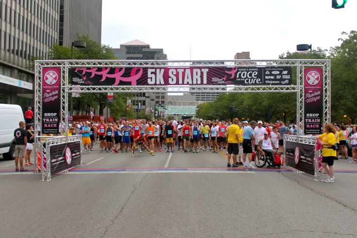 Runners waiting behind a 5K START banner on a scaffold gantry at a Susan G. Komen Race for the Cure, sponsor logos printed across the banner and the side towers