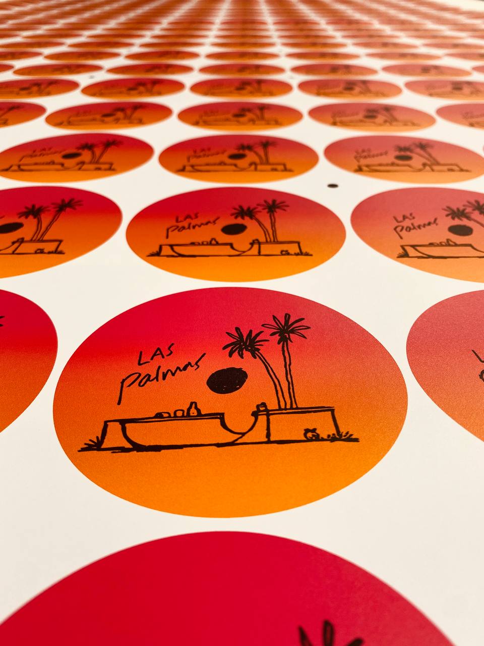 Freshly printed sheet of round Las Palmas stickers in an orange to red gradient, each with a line drawing of a poolside scene and palm trees