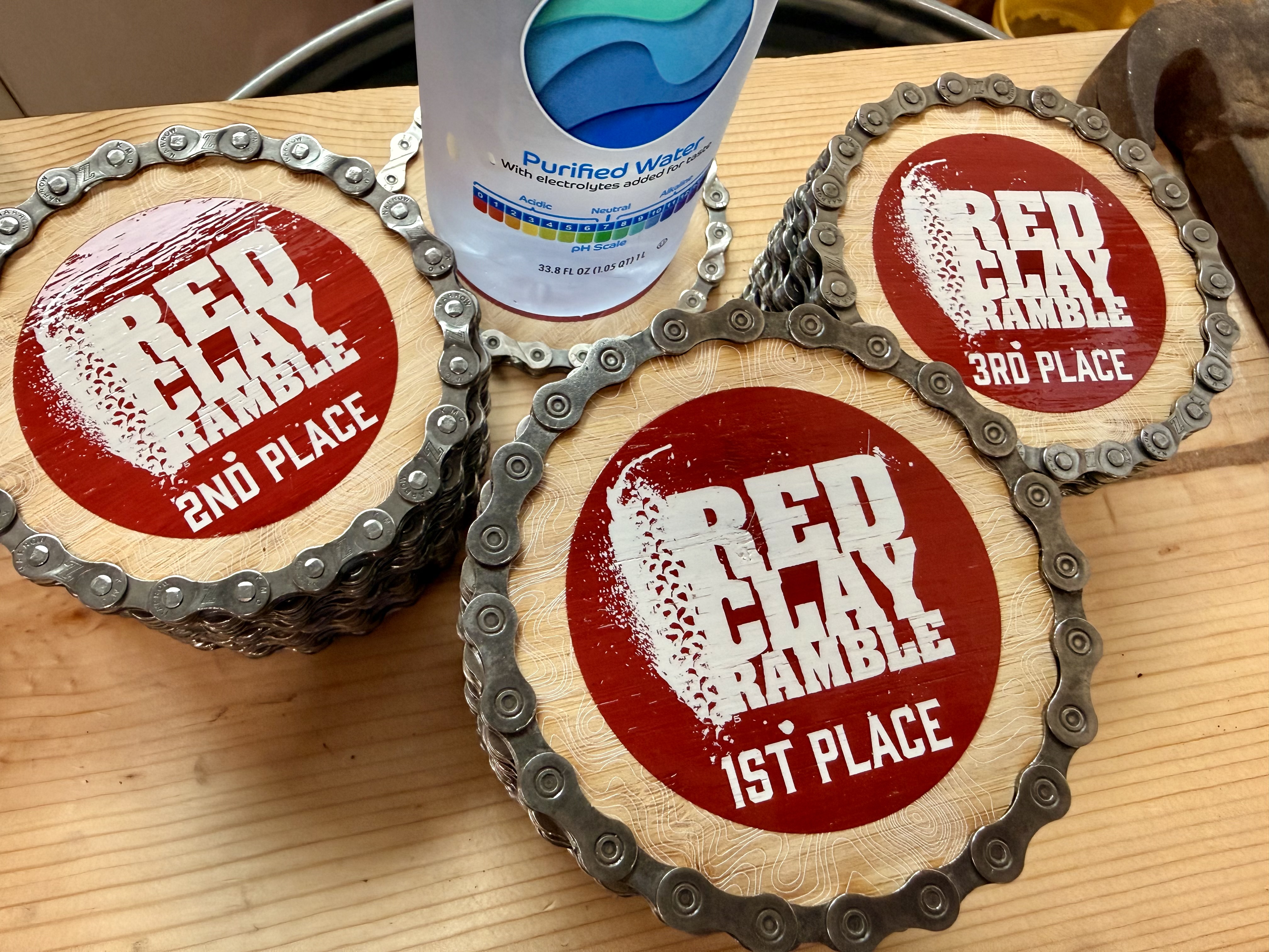 Three bamboo Red Clay Ramble coasters ringed with reclaimed bike chain, printed 1st, 2nd and 3rd place in red and white, on a workbench beside a bottle of water